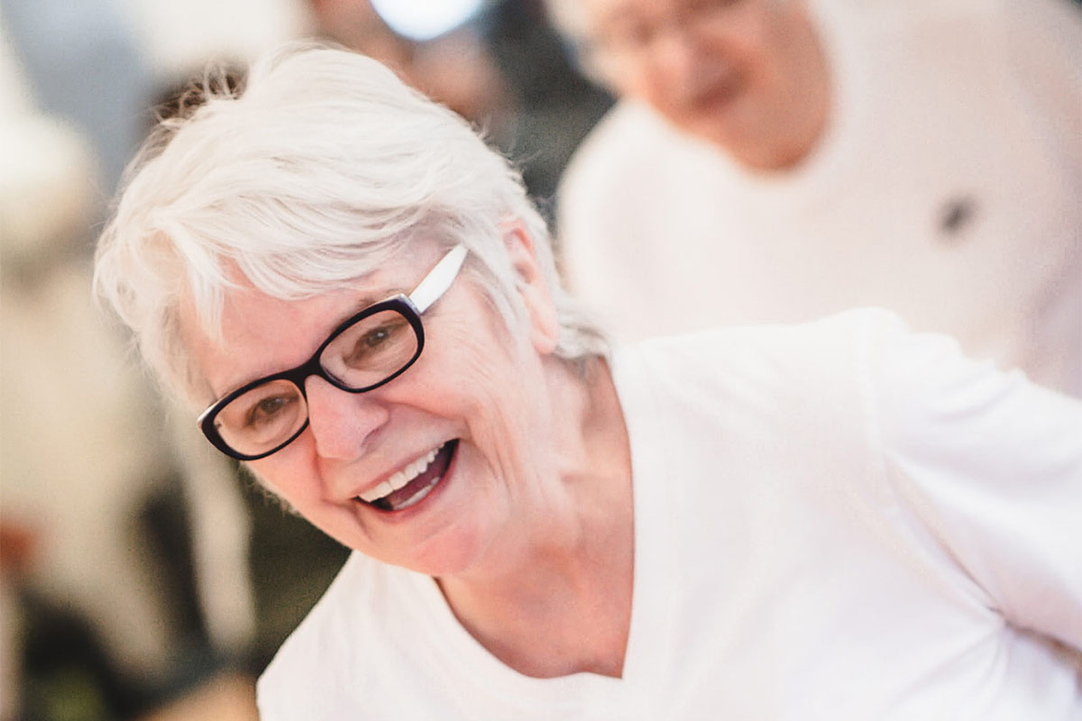 A female senior member in a white shirt and glasses laughs while jogging in place during a Senior Fitness class. Another senior member and a mirror are blurred in the background of the photograph. 