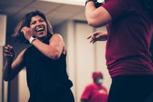Two women members dance together and laugh in a Cardio Funk class at Hays Communities YMCA. Focus of the photo is on woman in black shirt. 