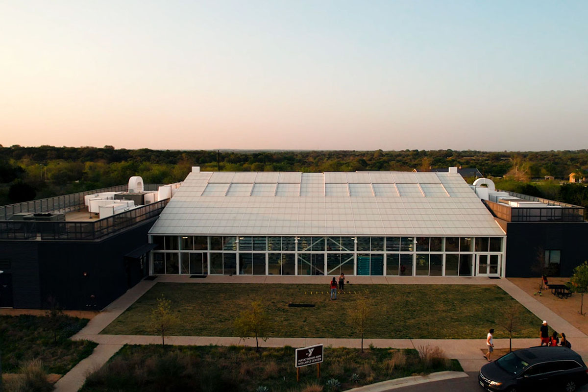 A modern-designed building with a natatorium in the center. Natatorium is white and lined with windows. Buildings on sides are grey and lined with metal. Grass lawn and parking lot sits in front. Behind building is the Camp Moody treeline.