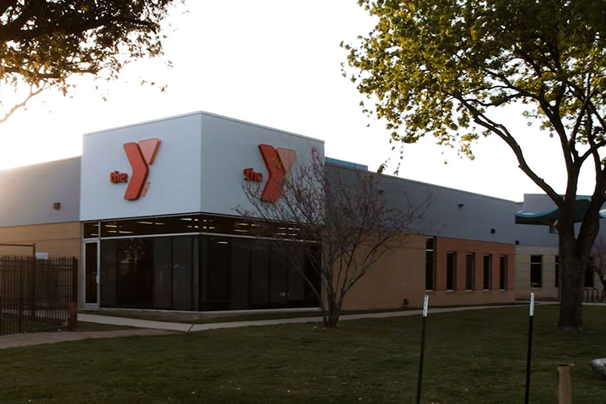 The corner of a building with floor-to-ceiling windows and a Y logo on two faces of the building. A metal fence is in the corner of the frame, and a sidewalk goes around the building.