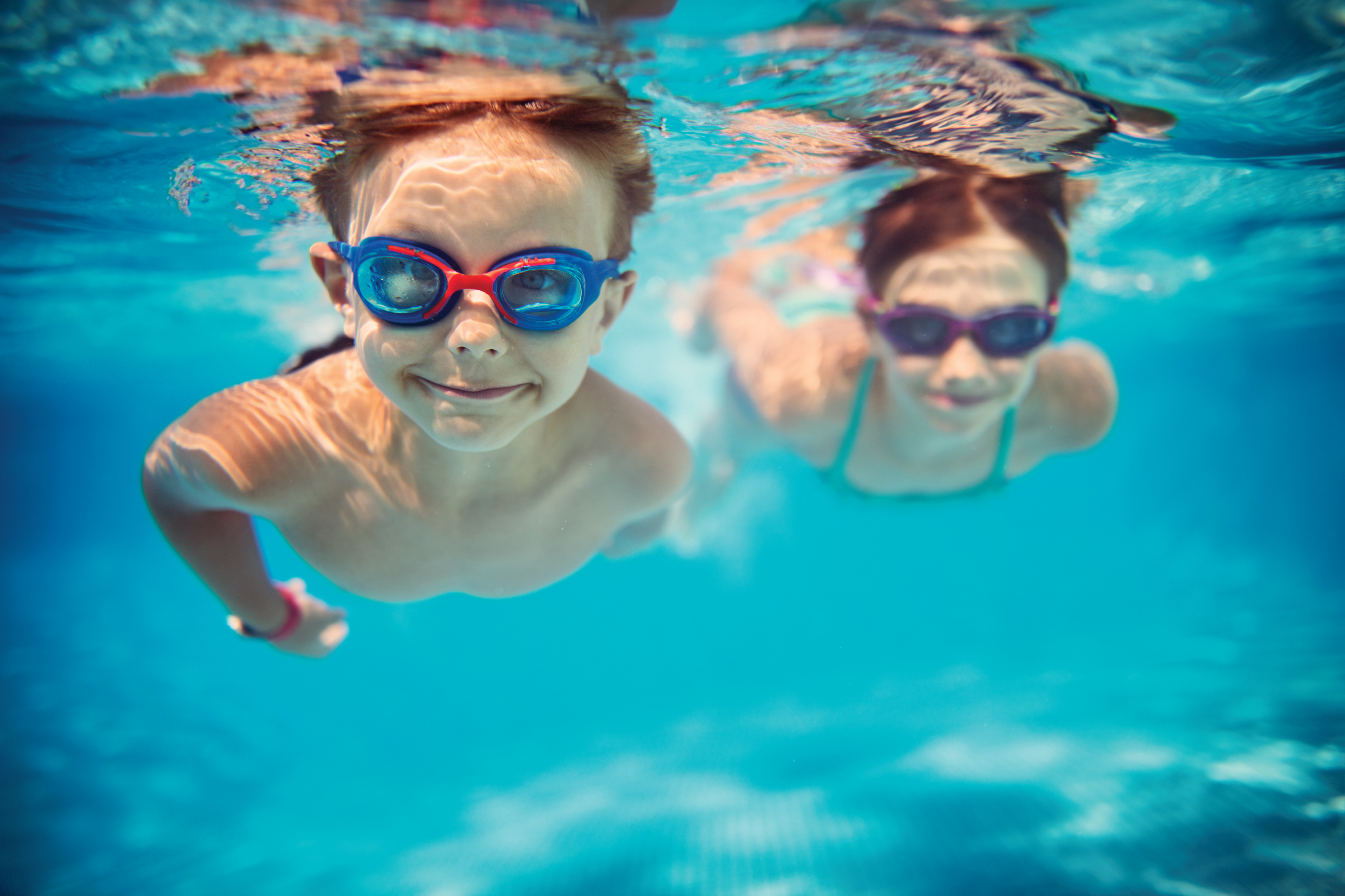 Two children swim under blue water towards the camera smiling.