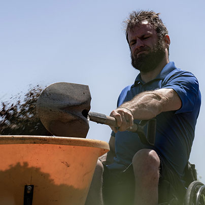 A bearded man wearing a blue polo shirt and using a wheelchair shovels dirt into an orange wheelbarrow. The dirt is in motion from the shovel to the barrow.