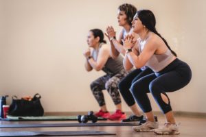 A group of 3 women in line complete of set of squats in their North Austin YMCA HIIT class. Focus of photo is on woman in foreground of photo. Yoga mats and dumbbells on floor in front of them. 