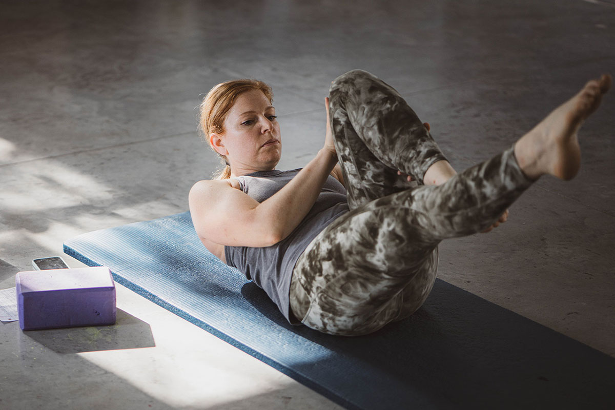 A woman lies on a yoga mat on a gray concrete floor with one leg extended. Sunlight streams through a window onto the woman’s shoulder and a yoga block