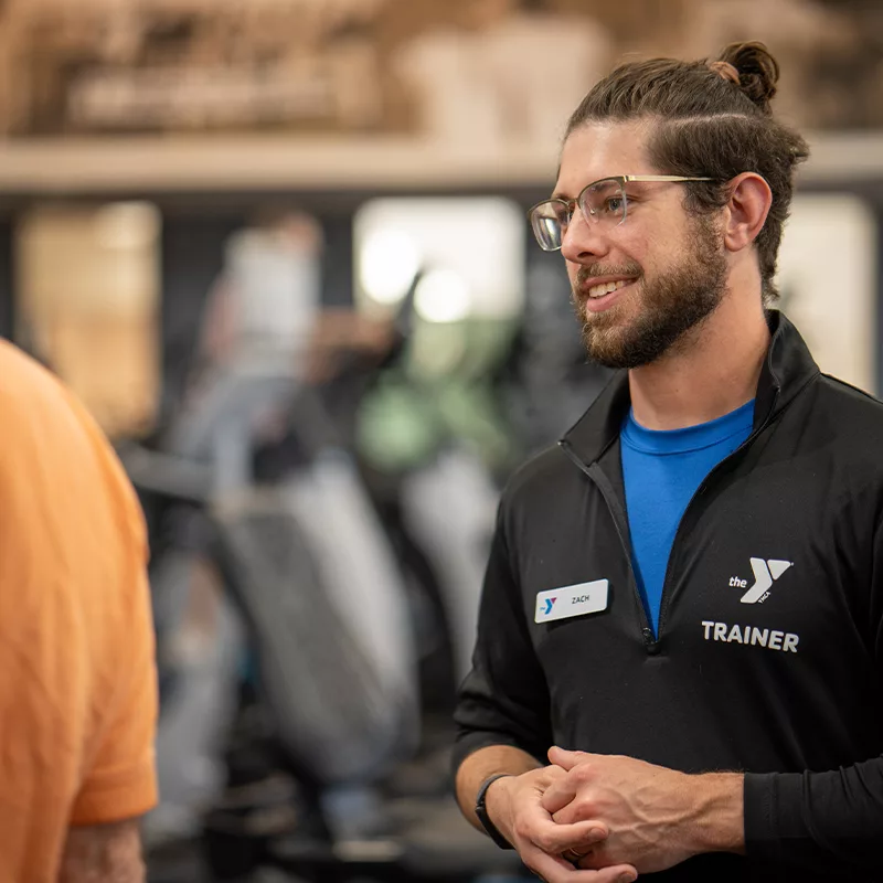A personal trainer smiles while helping a YMCA member with gym equipment.