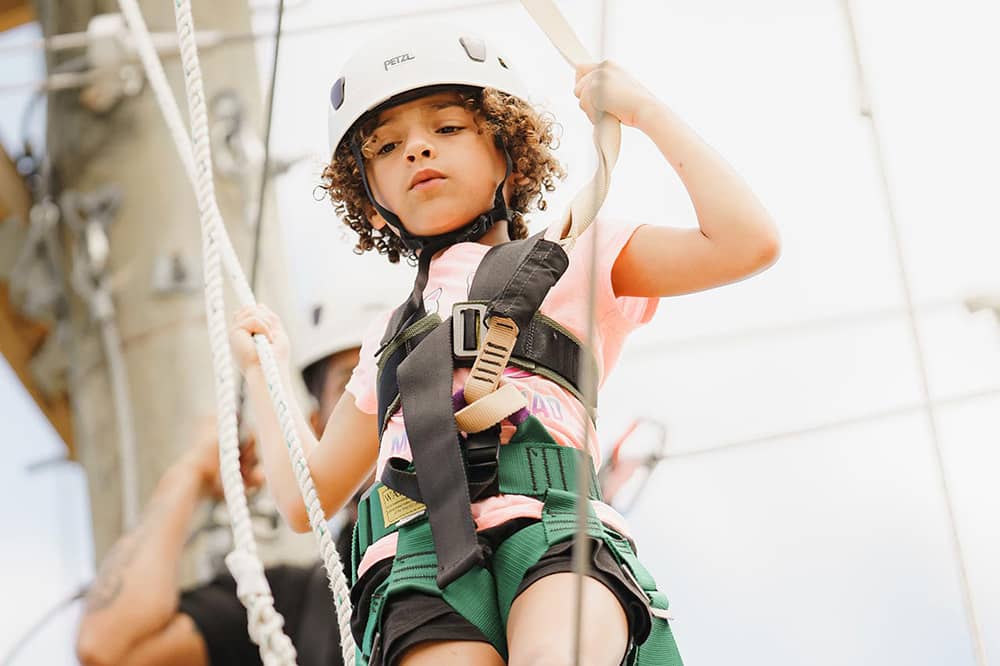 A young girl in a pink shirt and white helmet walks across the ropes course at YMCA Camp Moody, looking down below her. The ropes course and cloudy sky are blurred in the background of the photograph.
