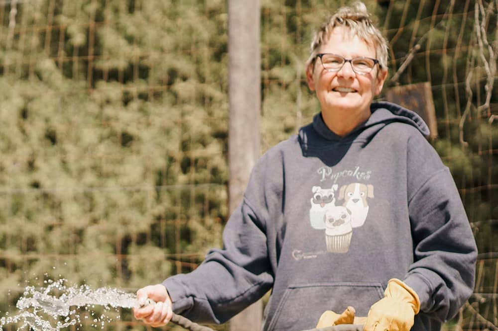 A senior female member in a blue hoodie and glasses smiles for the camera while watering a garden plot with a gray hose. A wire fence and treeline are blurred in the background of the photograph.