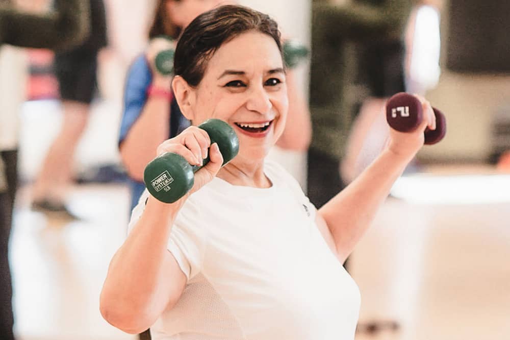 A female senior member in a white shirt and green and purple dumbbells in her hands smiles while doing a weight lifting exercise during a Senior Fitness class. Other senior members and a mirror are blurred in the background of the photograph.