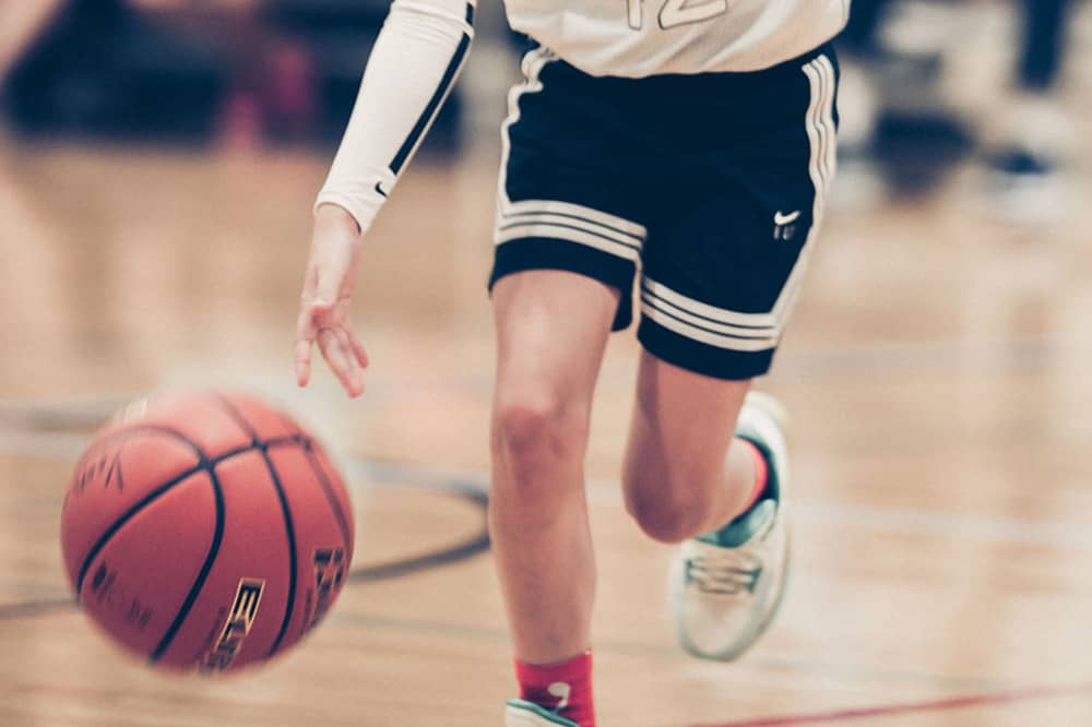 A girl in a white basketball uniform and black headband dribbles a basketball down the court. Parent spectators and the basketball court are blurred in the background of the photograph.