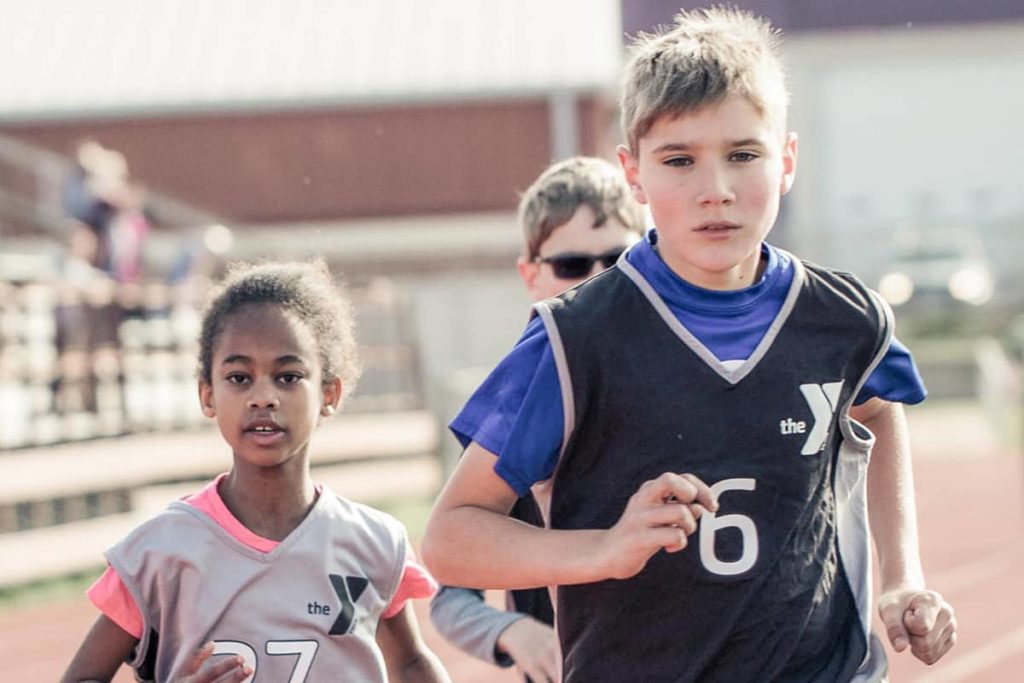 Two boys and one girl member dressed in black and gray track uniforms race against each other on a track. A track, football field, and school are in the background of the photograph.