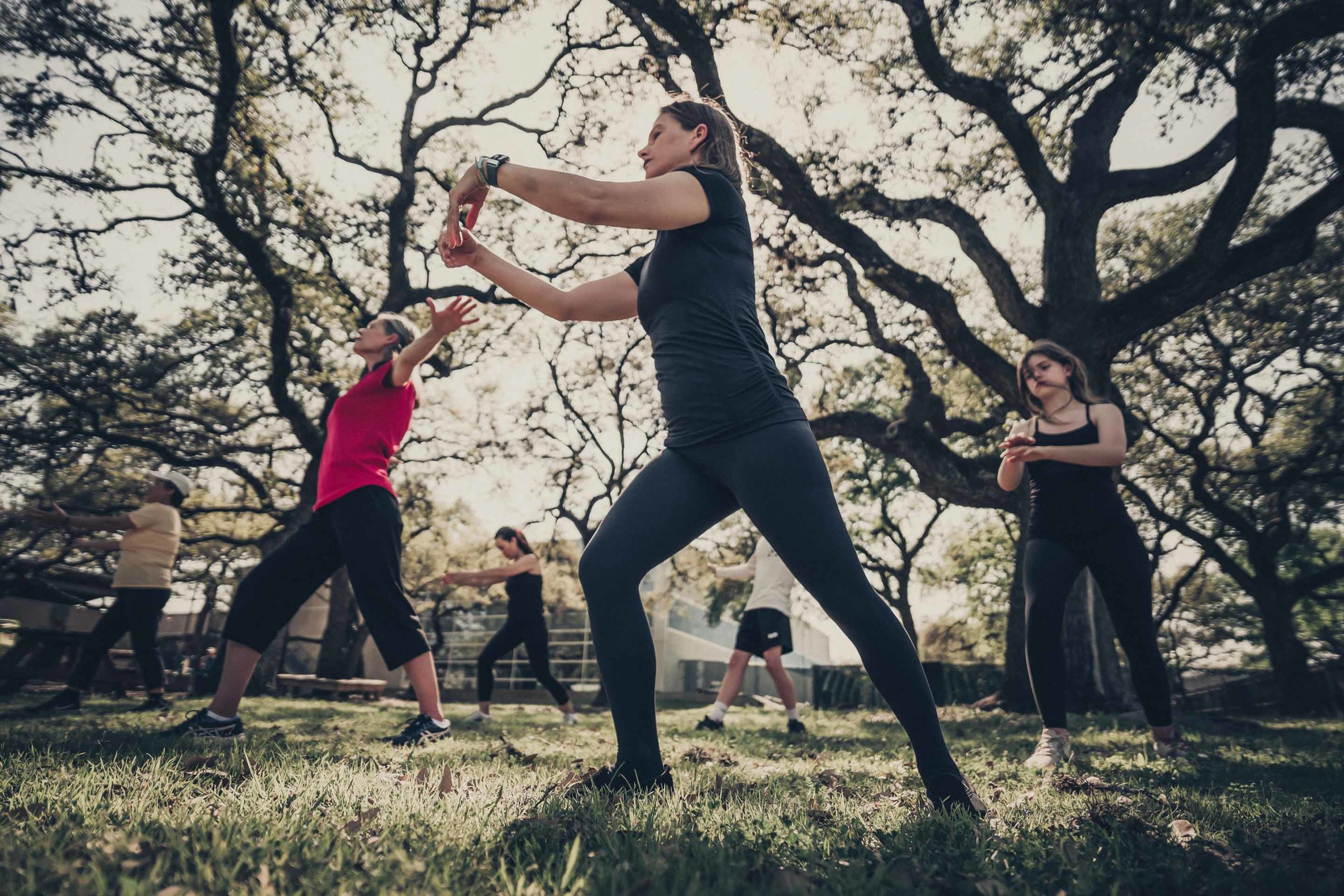 Woman member in black standing outside under tree canopy transitions into a Tai Chi pose peacefully. She is surrounded by other female members of various ages.