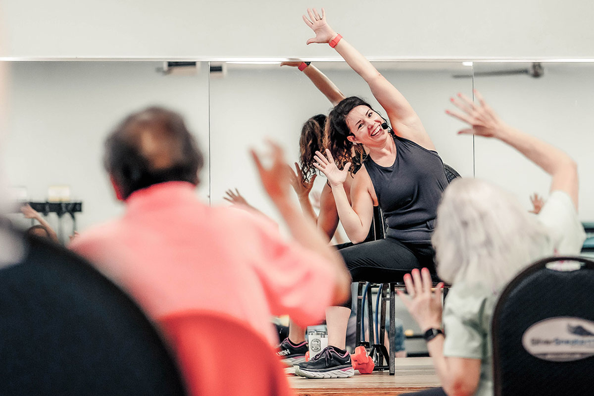 A group exercise instructor leads a class in seated movements. The instructor and the class participants have their arms raised over their heads. Everyone is leaning toward the left side of the photo.