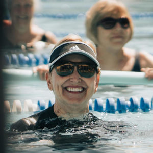 Women participate in an outdoor aqua cardio class. A woman wearing a black shirt, a black visor and sunglasses smiles directly at the camera. 