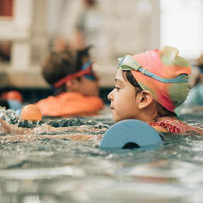 A child wearing a colorful pastel swim cap and goggles is in a pool. The child is using a foam flotation device.