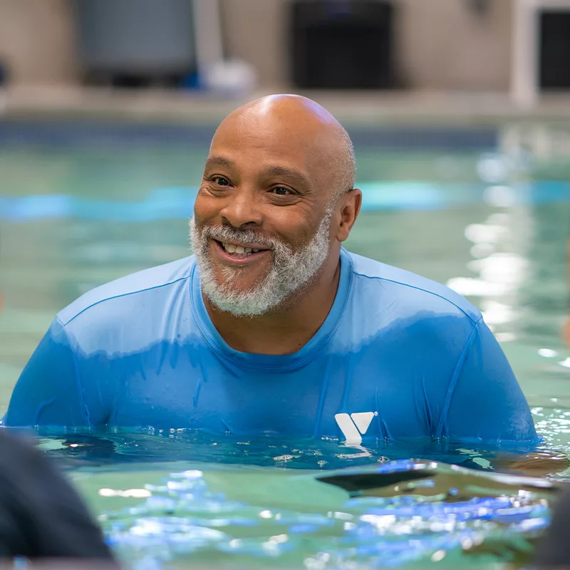 A YMCA employee smiles from a pool. He is guiding children who sit on the edge of the pool in swim instruction.