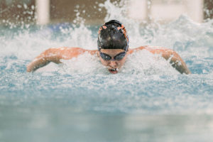 A young adult male swimmer in a black swim cap and goggles swims the butterfly stroke in a lap lane at YMCA Camp Moody. Pool lanes and the natatorium’s walls are blurred in the background of the photograph. 