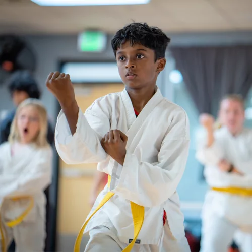 A child participates in a martial arts class. His fists are raised and he is wearing a white robe with a yellow belt.