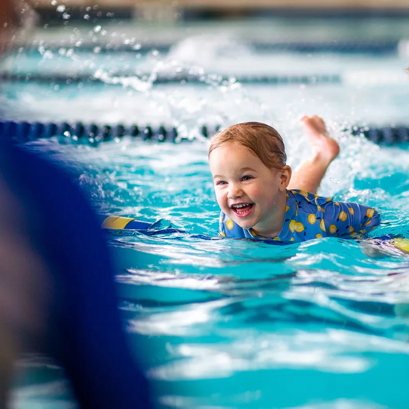 A young child smiles with delight while engaging in a swimming lesson