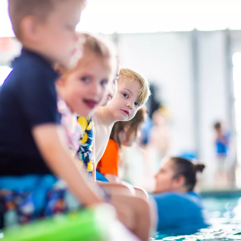 Young children sit at the edge of a pool waiting their turn for a swim instructor to include them in a lesson