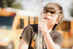 A boy in a black shirt and glasses looks and holds onto a long clear tube during an outdoor science experiment. A girl in a blue shirt, glasses, and a facemask watches on. A yellow school bus and white van are blurred in the background of the photograph. 