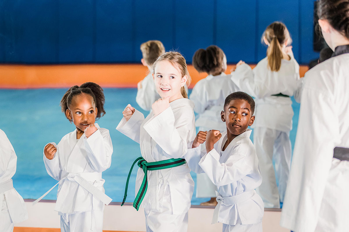 Three children members, two girls and one boy, in Taekwondo uniforms practice their forms in front of their female adult instructor. A wall mirror and blue floor mat are blurred in the background.