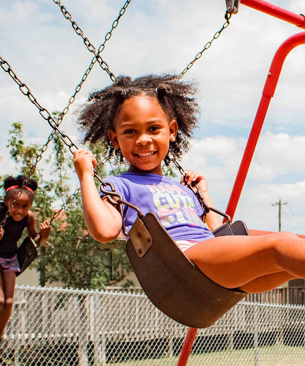 A girl in a purple shirt swings on a red swing set at a playground and smiles for the camera. Other kids swinging, a fence, and a yellow building are in the background of the photograph. 