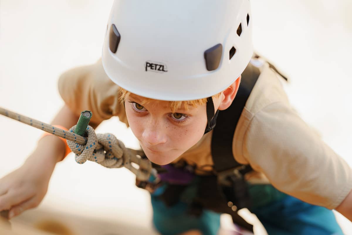 A boy in a tan shirt and white helmet climbs up the climbing wall at YMCA Camp Moody. A rope and harness are attached to the boy. The ground is blurred in the background of the photograph. 