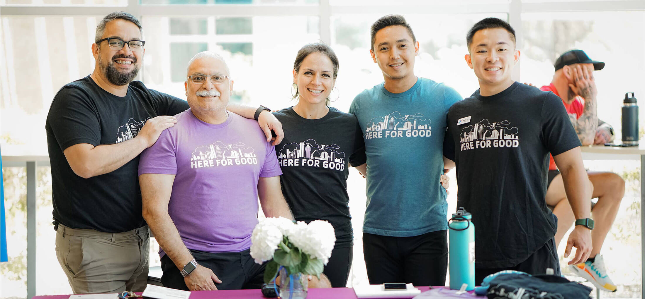 A group of five staff members wearing black, blue, and purple “Here For Good” shirts at the TownLake YMCA pose and smile for the camera while selling t-shirts for the 2022 Annual Giving Campaign. They are standing behind a table with flyers and t-shirts to sell.