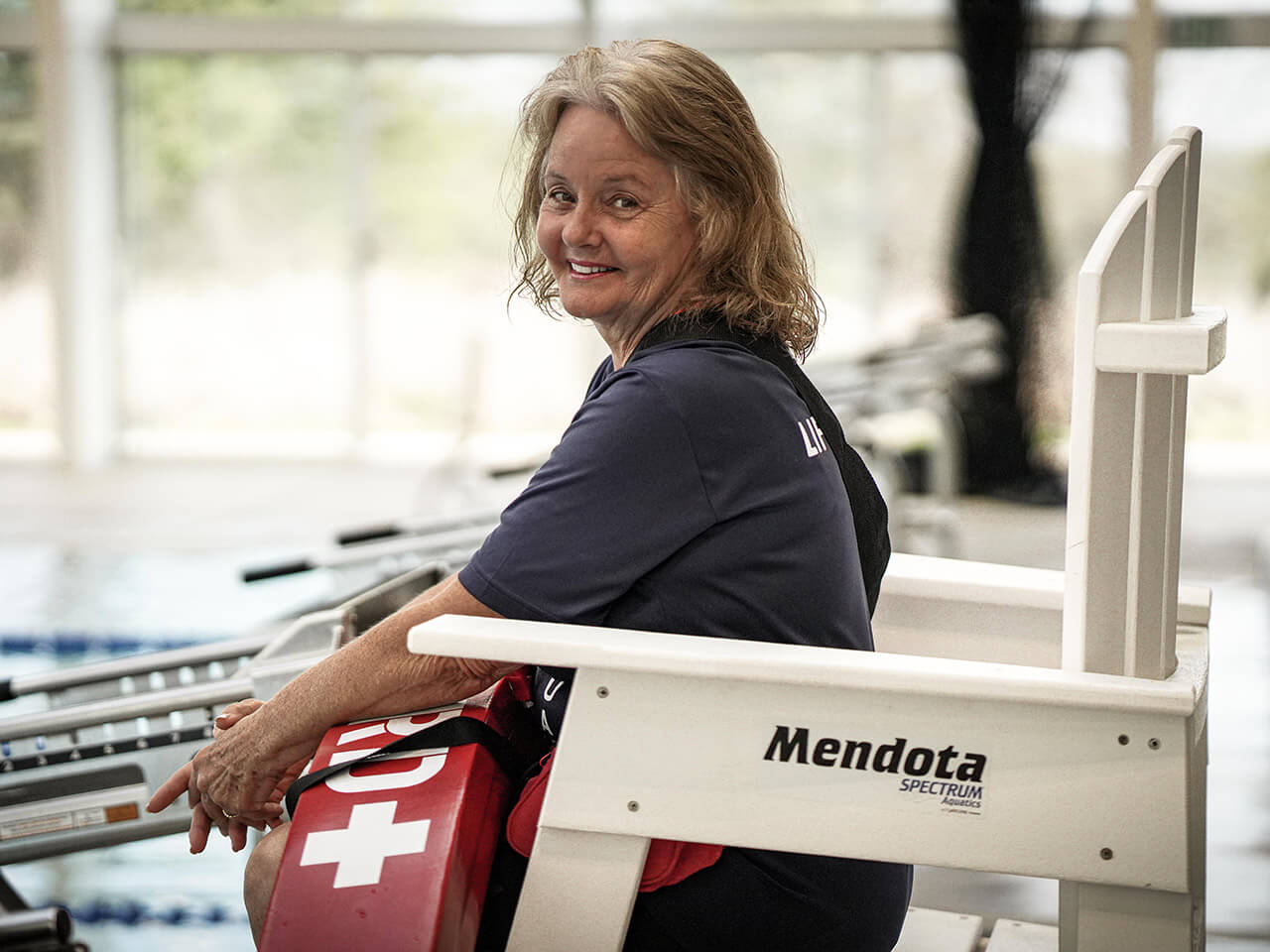 A lifeguard sits at a lifeguard stand holding a flotation device. She is looking over her shoulder and smiling.