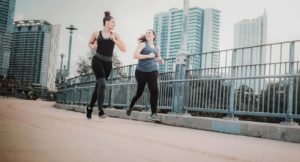 Two adult females in a black and gray sleeveless shirts smile and look at each other while running together in downtown Austin. The bridge they are running on and the Austin skyline are blurred in the background of the photograph.