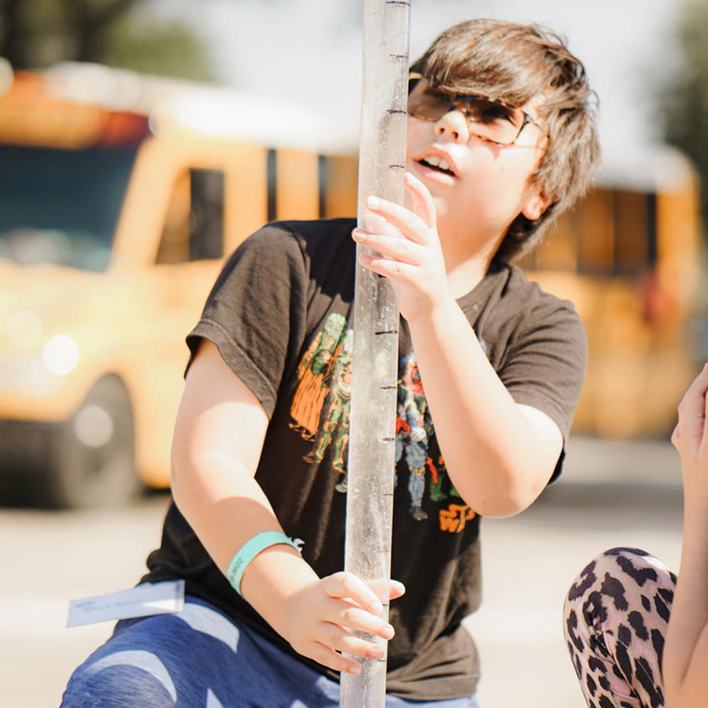 A boy in a black shirt and glasses looks and holds onto a long clear tube during an outdoor science experiment. A girl in a blue shirt, glasses, and a facemask watches on. A yellow school bus and white van are blurred in the background of the photograph. 
