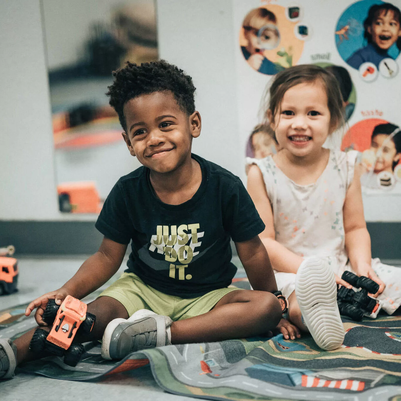 A little boy with a black shirt and gold shorts and a little girl with a white floral dress on the floor playing together. Poster in background with four children playing .