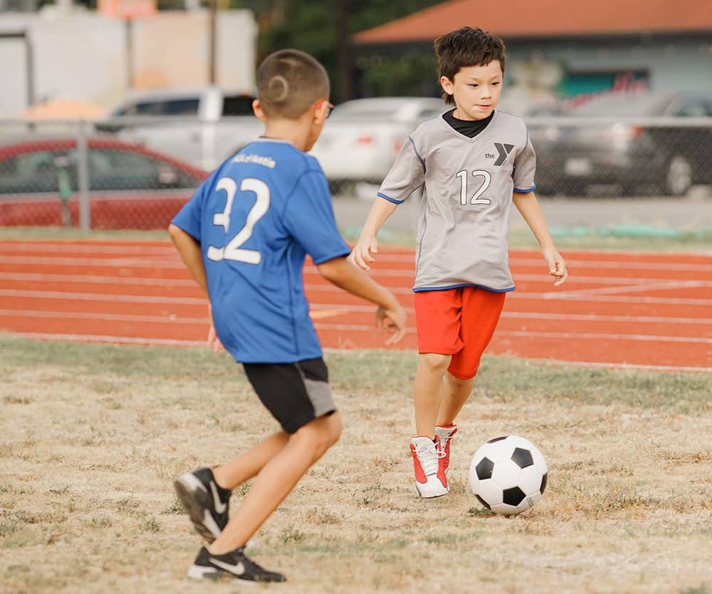 A boy in a gray soccer jersey kicks a soccer ball down the field while being defended by a boy in a blue soccer jersey and glasses. A track and neighboring business are blurred in the background of the photograph. 