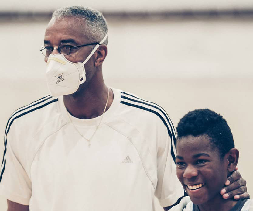 A male volunteer coach in a white shirt and facemask pats his son on the back after a basketball game. The boy is wearing a white basketball jersey. A basketball gym is blurred in the background of the photograph. 