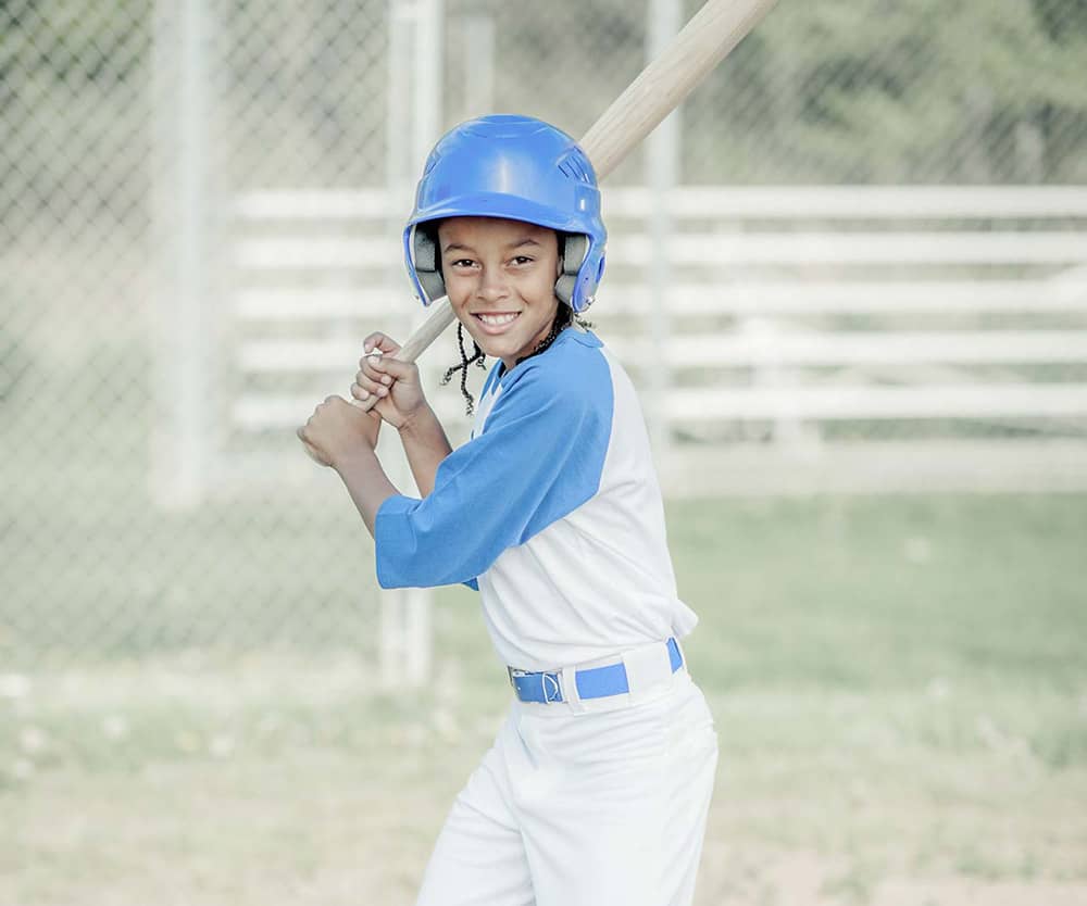A boy dressed in a blue and white baseball uniform with a bat in his hand stands at home plate and smiles for the camera. A dugout fence is blurred in the background of the photograph. 