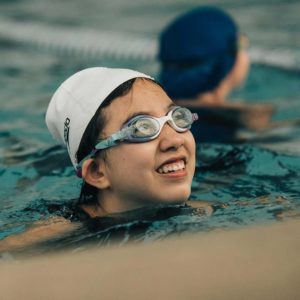 A girl in a white swim cap and goggles looks up at her swim instructor off camera as she treads water in a lap lane. Another child in a blue swim cap and lap lanes are blurred in the background of the photograph. 