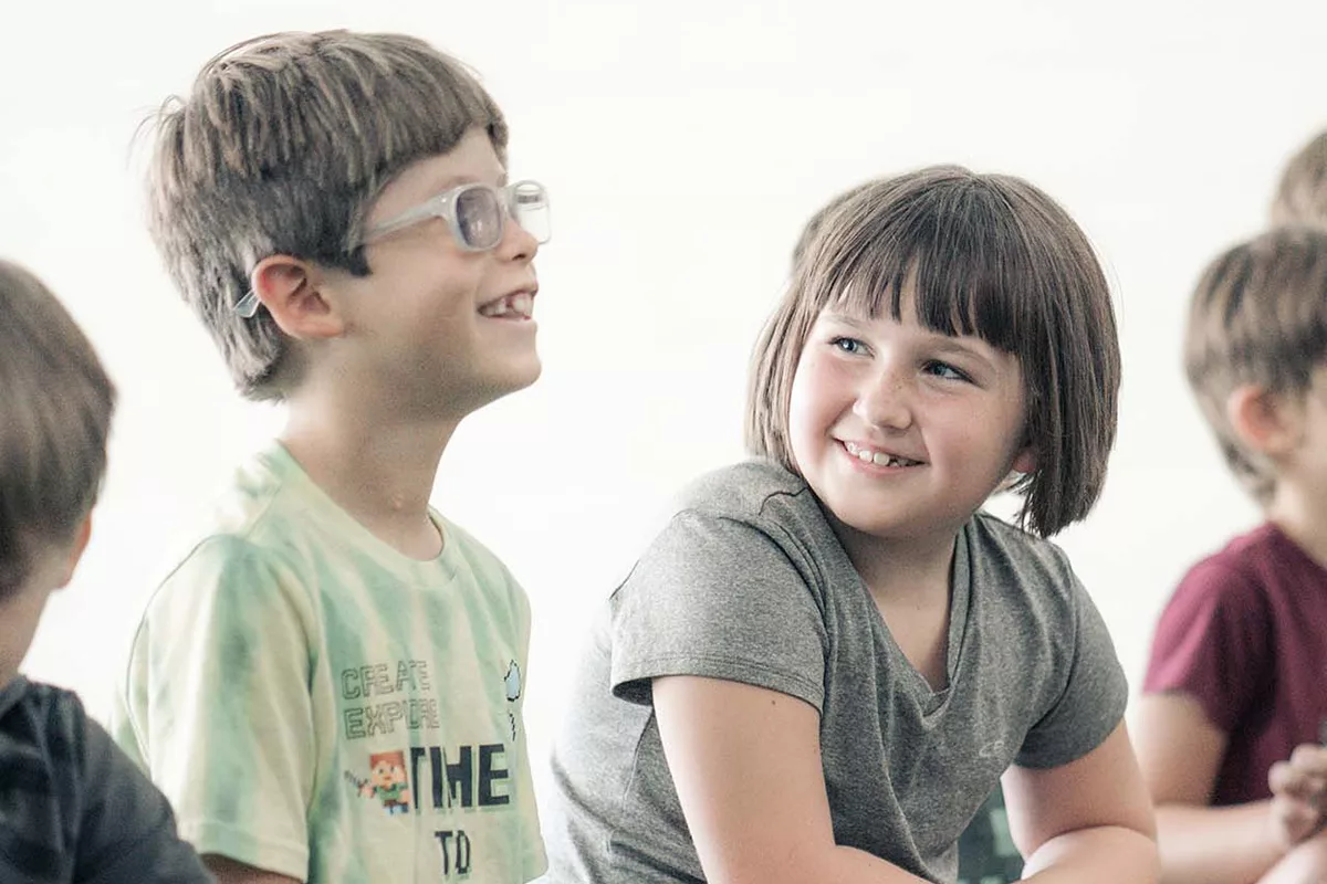 A girl in a gray shirt and a boy in a green shirt and glasses share a smile together while seated on the floor in a line with a group of other kids. The green floor and white walls of the room they are in are blurred in the background of the photograph.