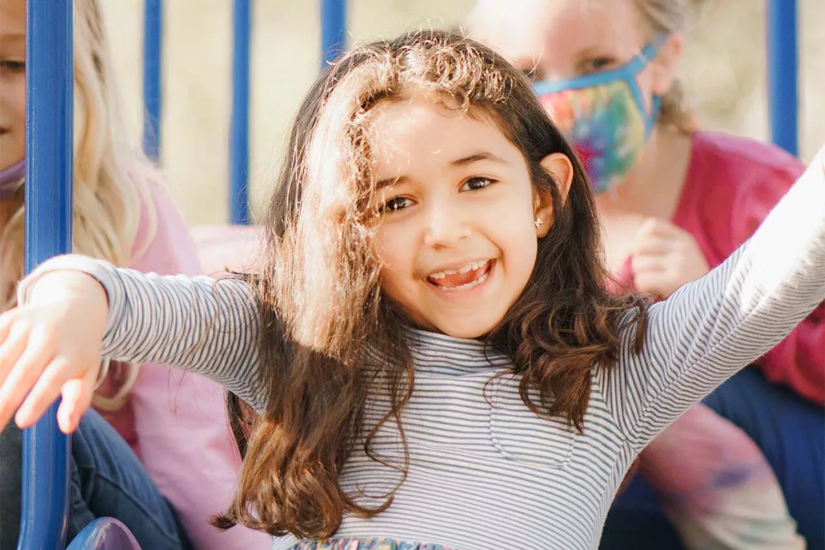 A little girl in striped dress cheers and smiles at the camera as she goes down a slide on a playground. Other girls in pink shirts and facemasks sit behind her at the top of the slide. Trees are blurred in the background of the photograph.