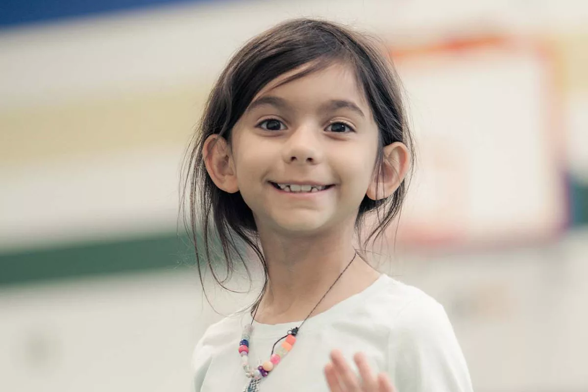 A girl in a green long sleeved shirt smiles and waves at the camera while playing tag in a school basketball gym. Other kids playing and the walls of the basketball gym are blurred in the background of the photograph.