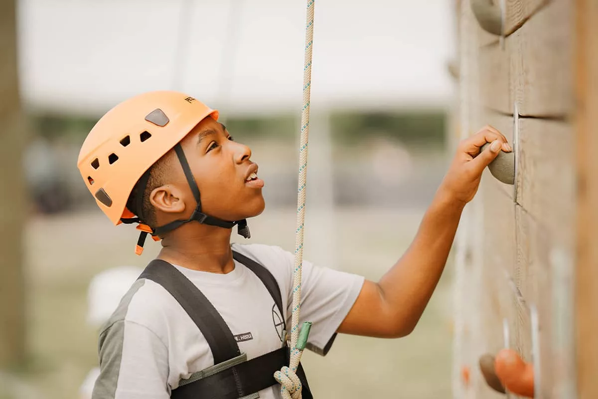 A boy in a gray shirt and orange helmet gazes up to the top of the climbing wall at YMCA Camp Moody as he grabs one of the wall pegs in front of him. A rope and harness are attached to the boy. The Camp Moody landscape is blurred in the background of the photograph. 