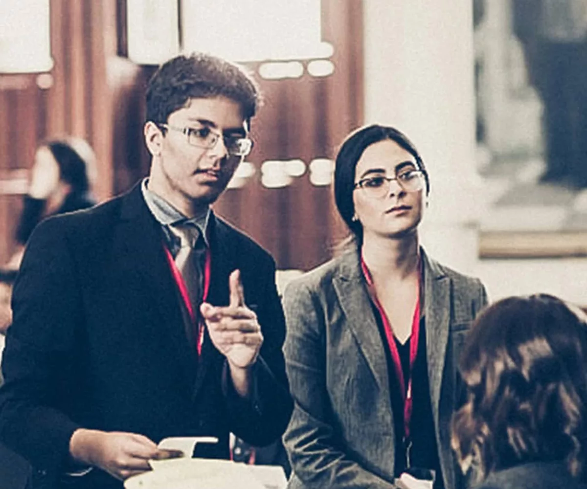 A male and YG teen dressed in business professional attire stands and speaks to a seated YG female teen in the Texas Capitol Senate Chamber. Other teens dressed in business professional attire seated at desks and the walls of the Texas Capitol Senate Chamber are blurred in the background of the photograph.