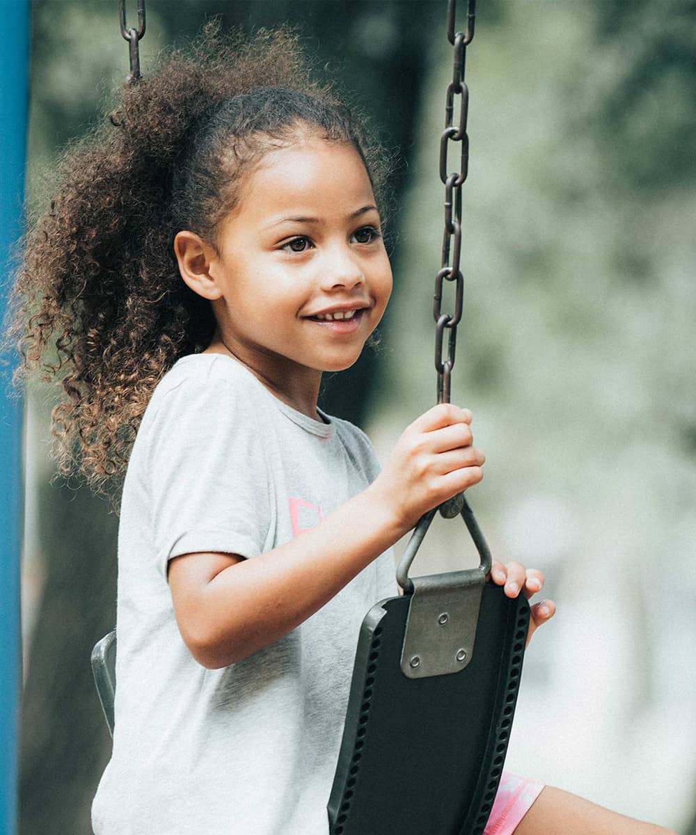 A little girl sits sideways on an outdoor swing. She is smiling.