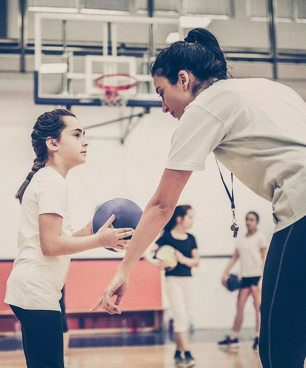 A girl in a white shirt holding a volleyball listens to her female adult volunteer coach in a white shirt give her instructions about how to properly serve a volleyball. Other teammates and a basketball gym are blurred out in the background of the photograph.