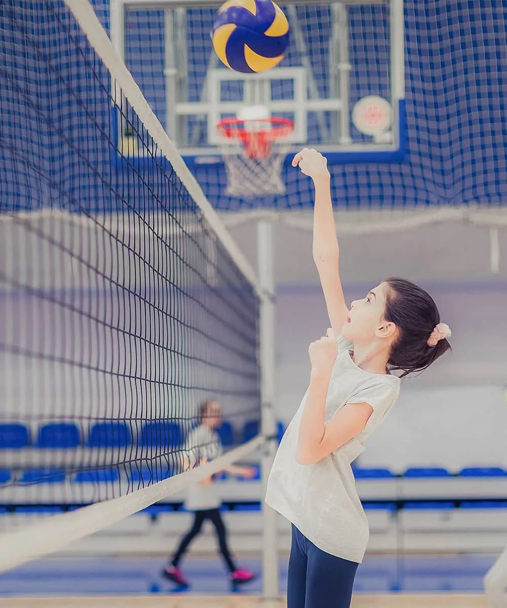 A girl in a white shirt serves a volleyball over the net in practice. Other teammates and a basketball gym are blurred out in the background of the photograph.
