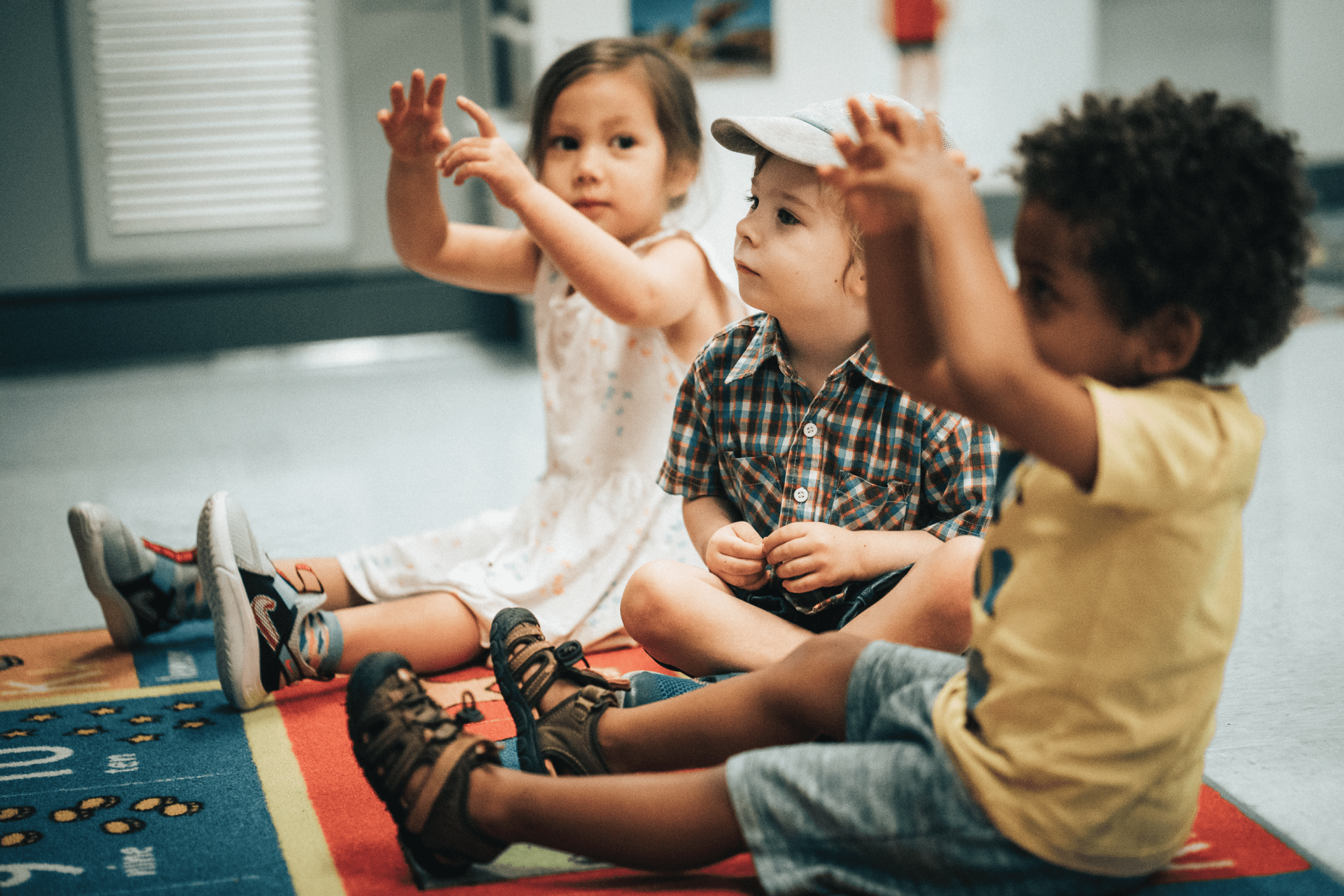 Children sit on a play mat and gesture with their hands.