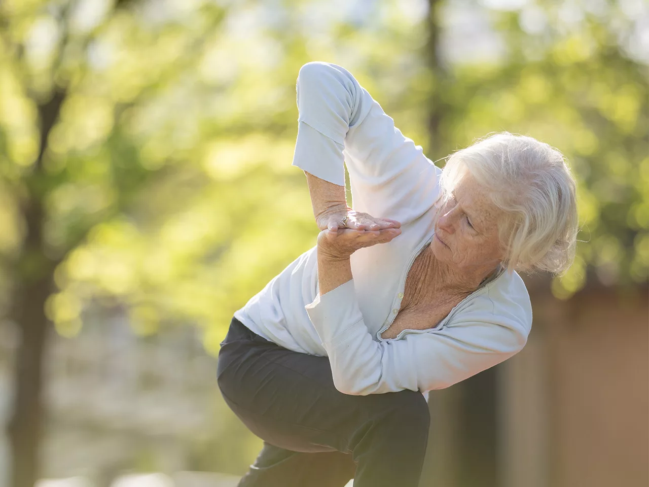 A woman bends at the waist and turns with her arms pressed together. She is outdoors in a green park.