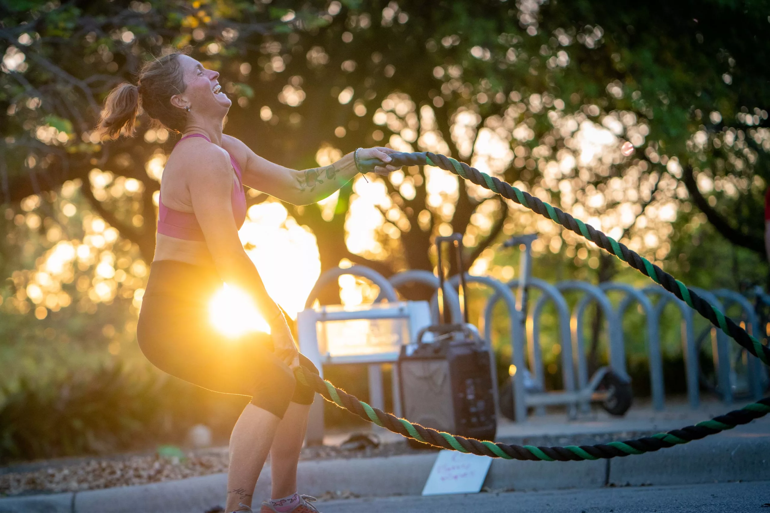 A woman exercises outdoors in the setting sun using battle ropes. Her head is flung back in joyful exertion.