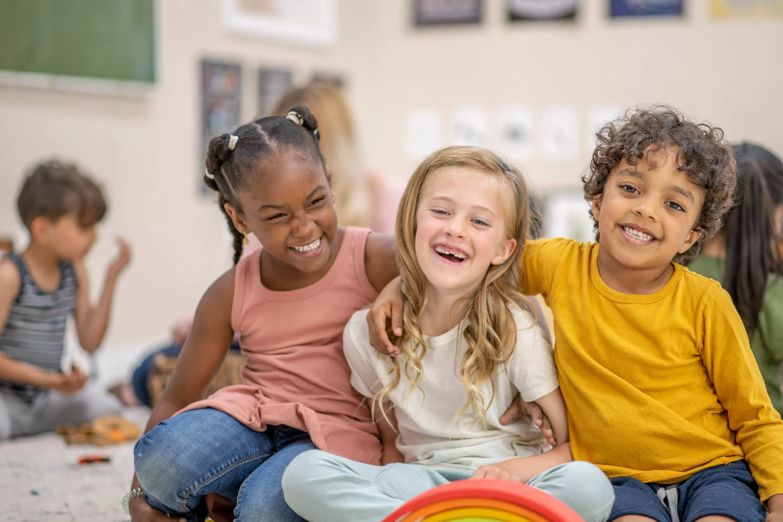 Three children sit in a colorful room. They have their arms around one another and are smiling.