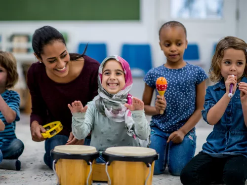 Kids in a classroom with a teacher hold a variety of musical instruments. A joyful child bangs the drums.