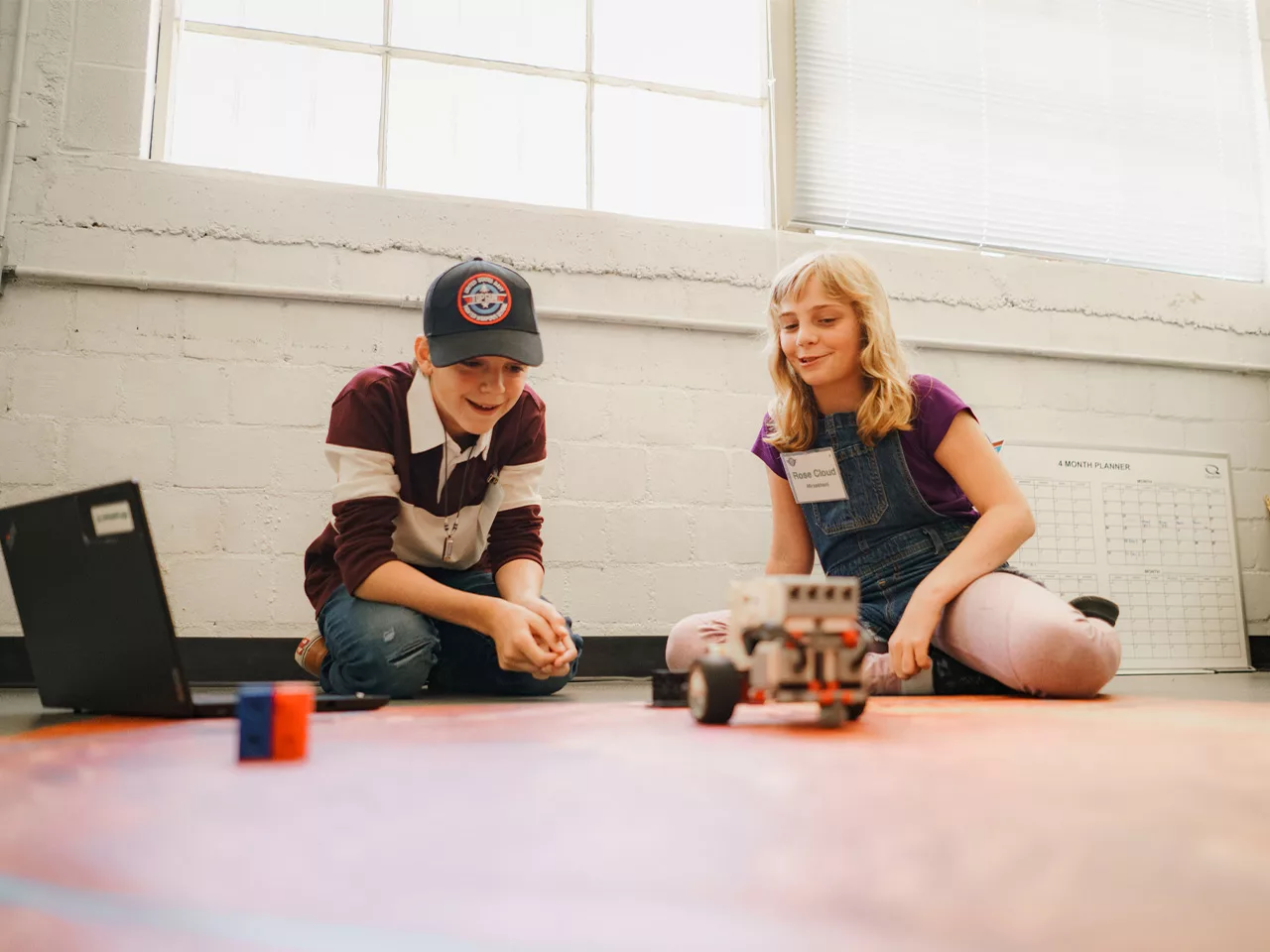 Two children sit on a floor operating a robot they have built.